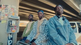 Three Black friends in street style clothes leaning on car hood, holding boombox and posing together at camera next to sticker-covered phone booth under urban overpass. Group portrait, zoom-in shot - Powered by Shutterstock - Get 15% off with code: PIKWIZARD15
