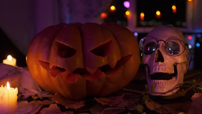 Halloween carved pumpkin next to skull and candles stands on doorstep of house among dry foliage burning candles in darkness at night party, background.