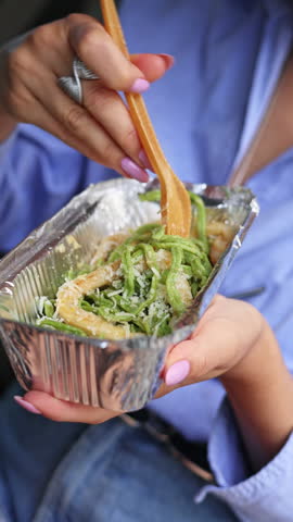 hungry woman eating green color pasta with seafood, sitting in car. lunch to go in box and girl in driver seat. lady in blue office shirt with short hair.