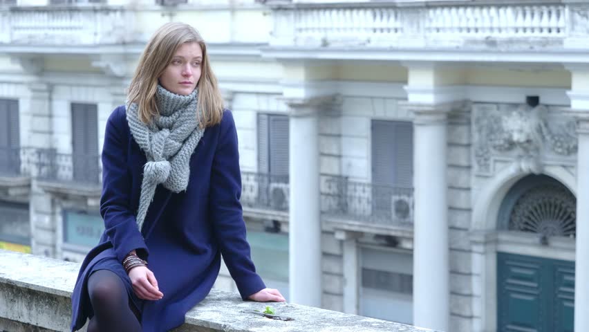A stunning young blonde woman sits on a city wall, gazing into the distance, lost in her thoughts and visibly unhappy