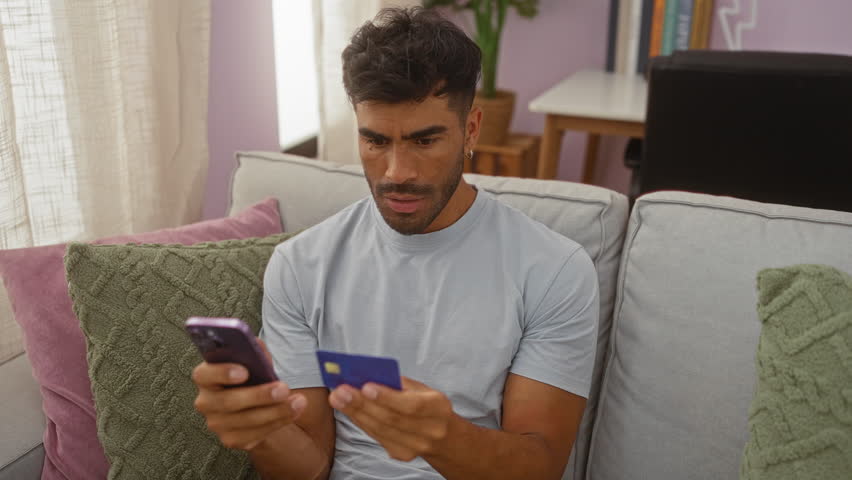Young man with dark hair sitting on a grey couch in a modern living room, holding a blue credit card and a mobile phone with a worried expression.