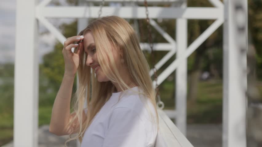 A blonde woman swings lightly in the park, her smile wide and her expression full of peace and happiness, enjoying a carefree day.