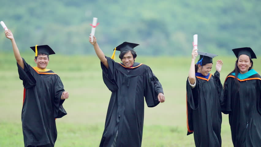 Four graduates are standing in a field, holding their graduation caps and gowns. They are all smiling and waving, celebrating their achievements
