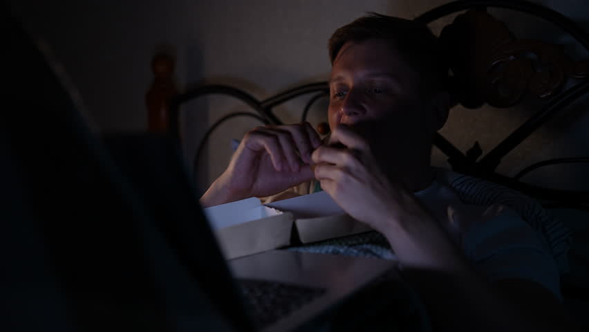 Hungry young man relaxing evening at home, lying comfortably in bed while watching movie on laptop and eating tasty fast food. Happy tired male enjoying eating burger in bed while using computer.