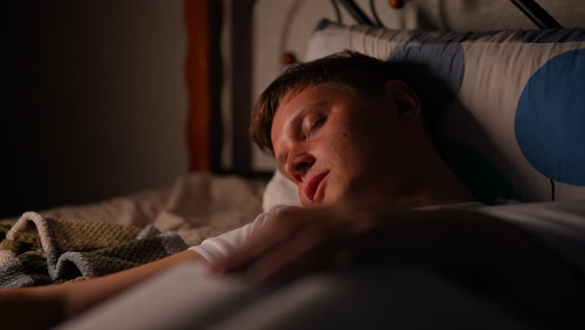 Closeup peaceful scene of young man sleeping peacefully in illuminated bedroom, radiating warmth and tranquility. Tired male sleeping soundly in bed, illuminated by warm light source in darkness room.