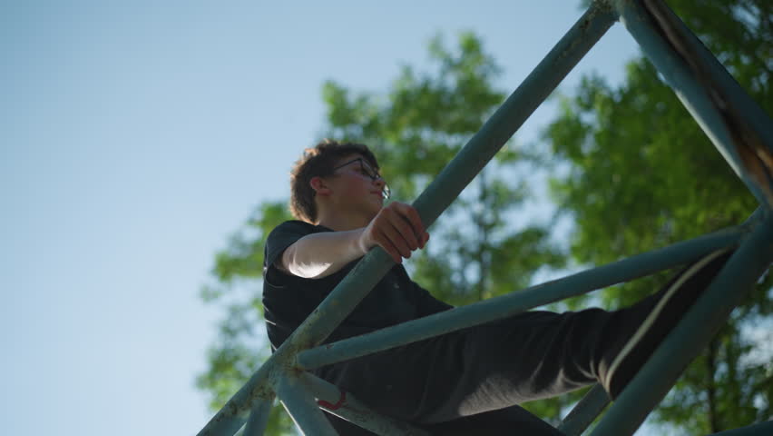 A young boy stands on an outdoor blue iron structure, looking thoughtful with both hands gripping the framework, a worn soccer ball is stuck within the iron bars