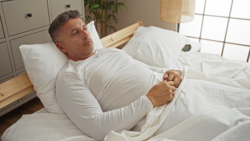 Middle-aged man sits in a comfortable bed in a bright, cozy bedroom, exuding a thoughtful expression while wearing a white long-sleeved shirt.