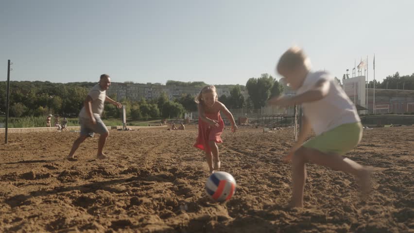 A happy family enjoys a sunny day playing volleyball on the beach. The young children run and laugh as their father joins in the fun, showcasing summer joy and family bonding.