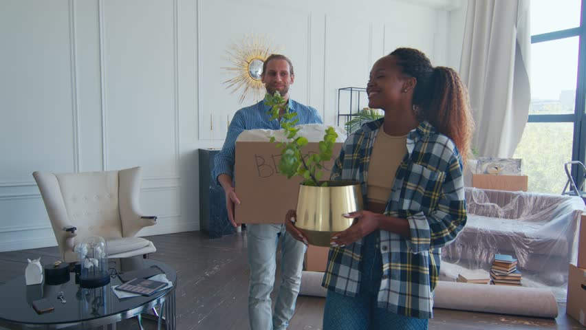 Young diverse couple from African American woman and Caucasian man carry box or home flower moving to new house and picking up personal belongings and walk through living room towards camera.