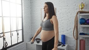 A young hispanic pregnant woman exercises with dumbbells in an indoor gym setting, showcasing her dedication to fitness and a healthy pregnancy amidst the supportive sport center environment. - Powered by Shutterstock - Get 15% off with code: PIKWIZARD15