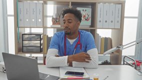 A young, african american man with a beard and stethoscope sits stressed at a desk in a veterinary clinic with a laptop, smartphone, documents, and a colorful animal poster in the background. - Powered by Shutterstock - Get 15% off with code: PIKWIZARD15