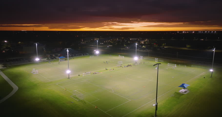 Aerial view of public sports park with children engaged in football game on grass stadium at night. Active way of life concept