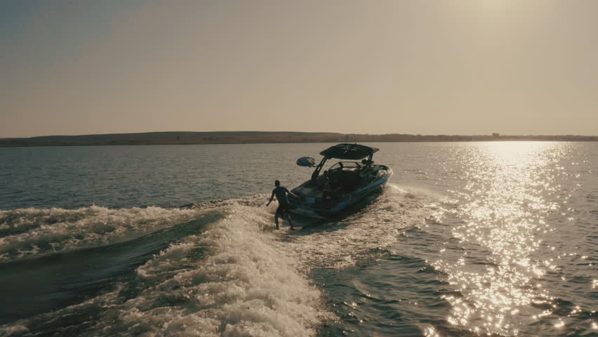 Flying over a wakeboarder surfing behind a boat. Aerial view of wakeboarding on the lake at sunset. Active recreation on the water