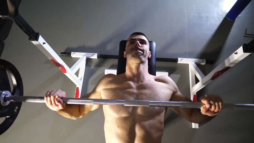 Strong sporty man doing bench press workout with a barbell at modern gym. Young muscular sportsman doing weightlifting exercises at sport club. Athletic guy training in fitness centre. Top view