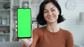 Happy young woman showing mobile phone with green screen sitting on sofa in living room at home. Color key, vertical template layout for advertising. Smiling female looking camera holding smartphone - Powered by Shutterstock - Get 15% off with code: PIKWIZARD15
