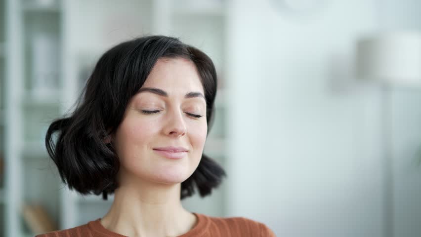 Young adult woman with closed eyes enjoying relaxation and tranquility in cozy living room on sofa at home. Calm and peaceful expression reflects a sense of mindfulness and inner peace. Close up