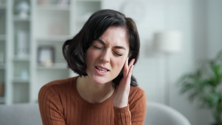 Earache. Sick young woman suffers from ear pain sitting on sofa in living room at home. Upset woman touches her ear with her fingers. She has furuncle, shingles, eczema, ear diseases or otitis media
