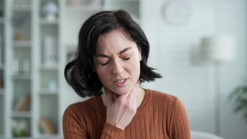 Sick young adult woman suffering from a sore throat while sitting in the living room at home. Upset female holding her neck in discomfort and pain, has first symptoms of cold, virus or flu. Close up
