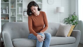 Young adult female suffers from painful severe knee joint pain while sitting on sofa in living room at home. Upset woman massages leg muscles, could not walk, illness, cramps or rheumatism - Powered by Shutterstock - Get 15% off with code: PIKWIZARD15
