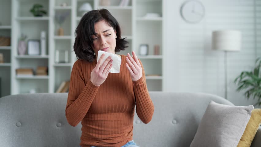 Young woman experiencing sneeze at home, sitting on sofa with tissue in hand. Upset female feels discomfort from allergies or cold in living room setting. Sick woman wipes her nose with a handkerchief