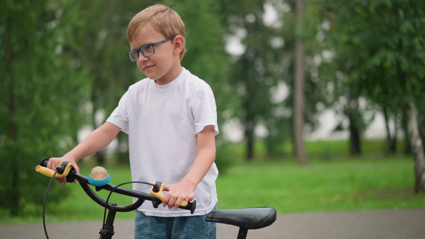 A young boy wearing glasses carefully observes his bicycle, holding onto the handlebars with a focused expression, he is dressed in a white shirt and denim shorts