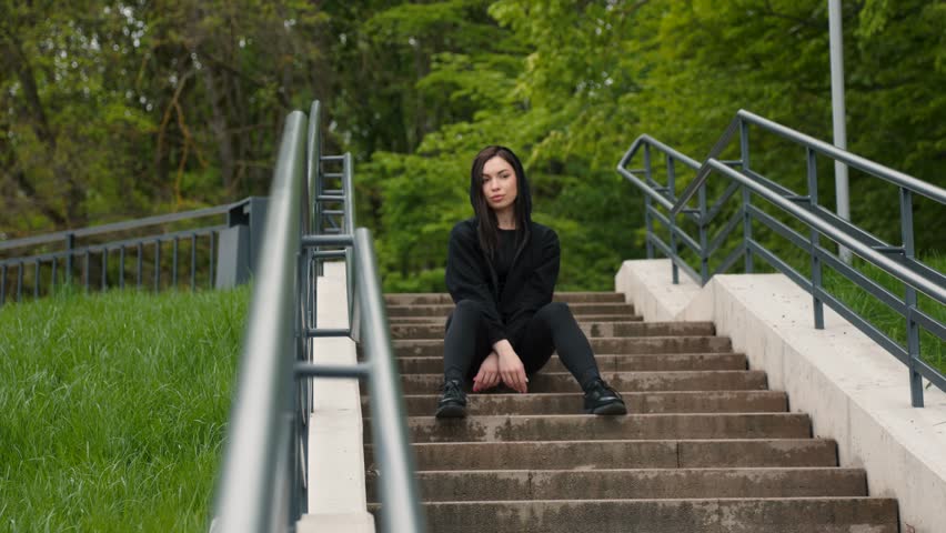 Attractive dark-haired sporty girl in a hoodie sitting outdoors in the park on the stairs. slow motion