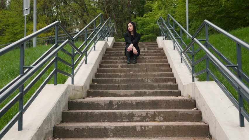 Attractive dark-haired sporty girl sitting outdoors in park on stairs. The girl looks to the side, then looks directly into the camera. slow motion