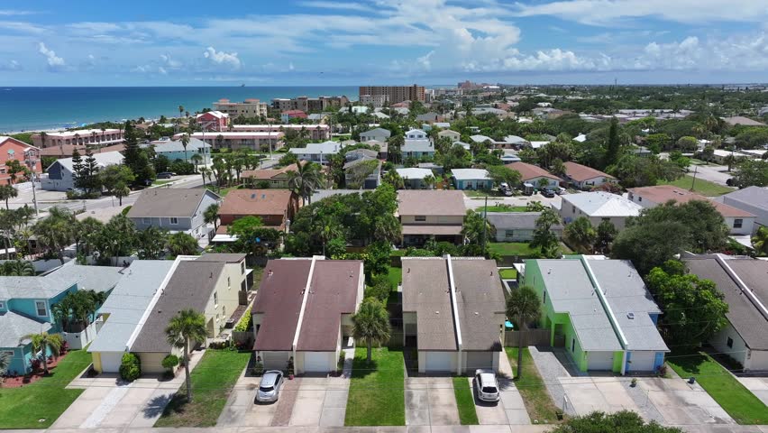 Rows of coastal homes leading towards the beach with ocean views. Suburban streets lined with houses and palm trees. Aerial view of Cocoa Beach, Florida, showing residential and beachfront properties.