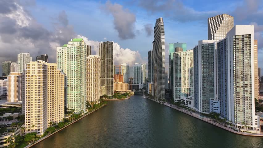 Brickell skyline in downtown Miami along the Miami River. High-rise condos and office buildings line the waterfront, creating a canyon-like urban landscape. Iconic towers reflect sunlight. Aerial.