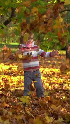 Happy girl laughs and throws autumn leaves in evening park at sunset. Child plays outdoors. Leisure weekend in nature. Vertical shot