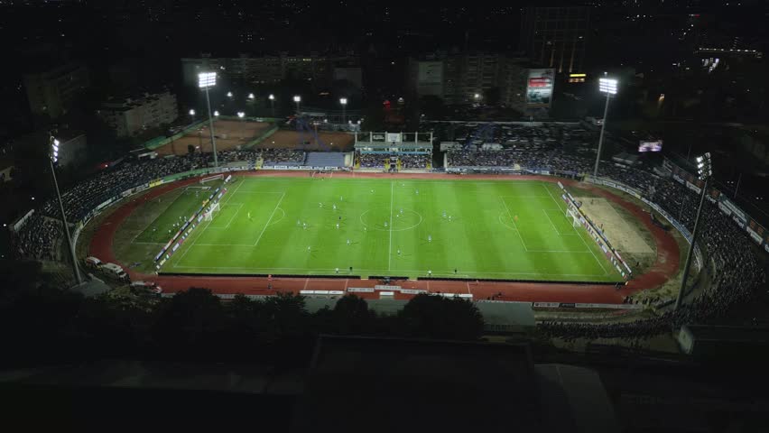 Drone footage capturing a night football match in a brightly lit stadium. Spectators fill the stands as teams compete on the field, creating an electric atmosphere in the city
