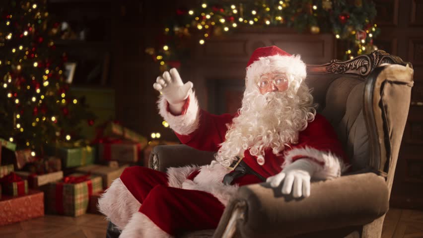 Santa Claus telling fairytales, congratulations and greeting, portrait in home. Old man in Santa costume talking to camera, sitting alone in armchair in cozy decorated room in Xmas eve, slow motion