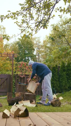 A middle-aged man is chopping firewood in the backyard of his house in early autumn