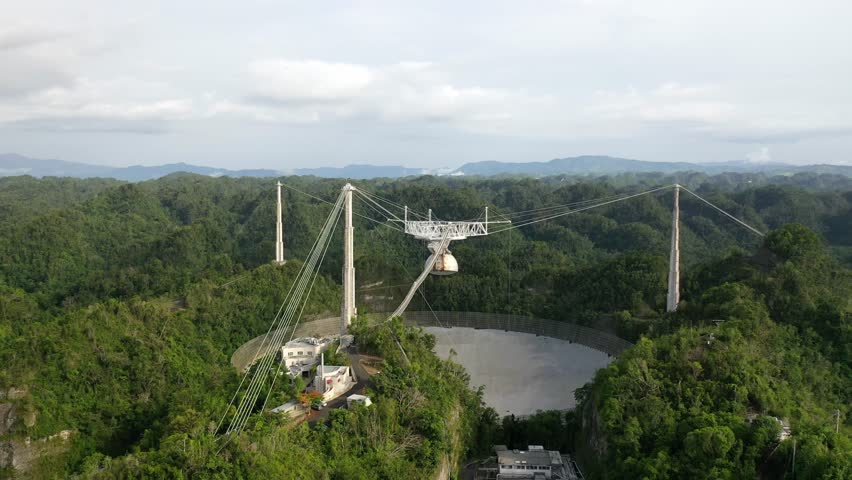 A drone shot of the Arecibo observatory radio telescope surrounded by green lush forests, with clouds background in Puerto Rico