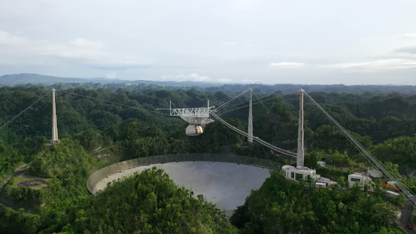 A drone shot of the Arecibo observatory radio telescope surrounded by green lush forests, with clouds background in Puerto Rico