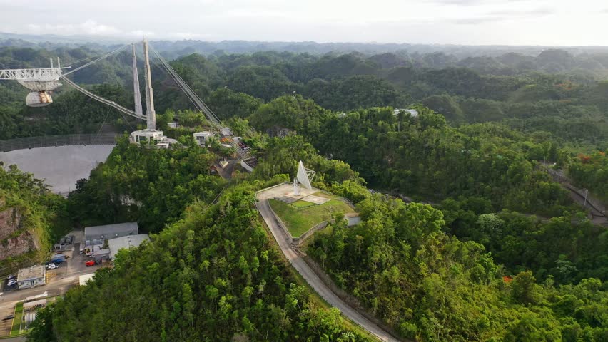 A drone view of Arecibo observatory radio telescope surrounded by green lush forests with road in the foreground on a hill in Puerto Rico