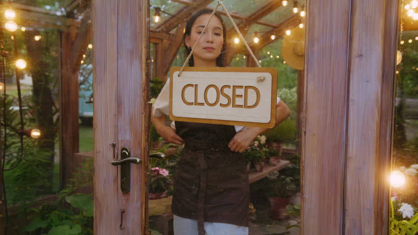 Attentive asian female looking aside before fixing welcoming sign and opening door with friendly smile. Happy floral shop owner preparing for new day in retail and feeling ready providing service.