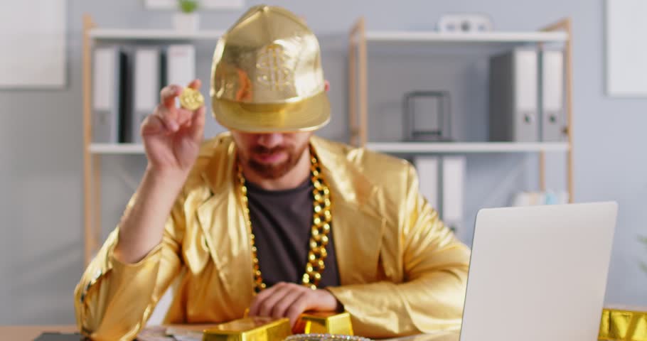 Successful rich happy man holding bitcoin coin cryptocurrency, wearing shining golden jacket, massive chain around neck, trading on stock market, investing Bitcoin cryptocurrency at home using laptop 