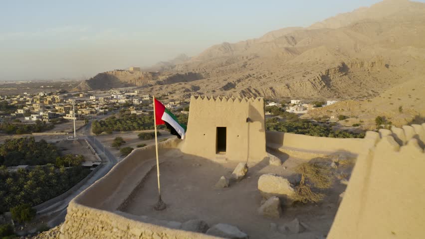An aerial at sunset of Dhayah Fort in Ras Al-Khaimah in the UAE, with the Emirates flag in full focus, revealing the town below