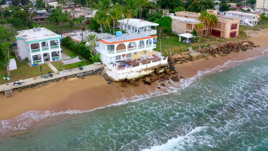 A drone shot of the Atlantic Ocean waves crashing on the rocks under a coastal house on sandy shore in Stella, Rincon, Puerto Rico