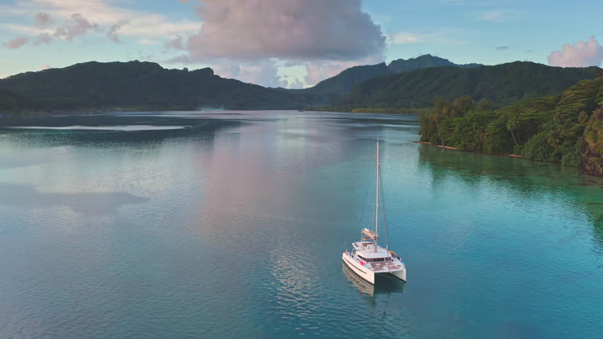 Aerial view catamaran sailing calm turquoise water lagoon near tropical island with lush green mountains and a beautiful sunset in background. Remote wild nature paradise, exotic summer luxury travel