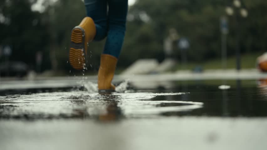 Close up a happy teenage girl in a yellow jacket and rubber boots runs through a puddle splashing water to the sides after rain in the park