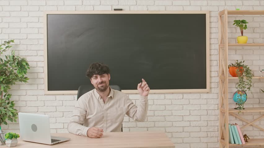 A male teacher sits at a desk in a classroom with a blackboard in the background and points upward with his index finger. Workspace, copy space.