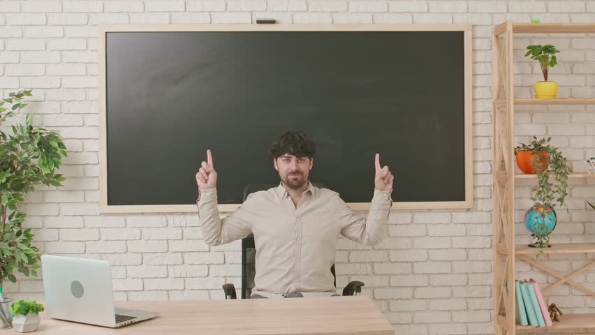 A male teacher sits at a desk in a classroom with a blackboard in the background and points upward with his index fingers. Workspace, copy space.