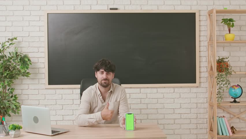 A male teacher sits at a desk in a classroom in front of a blackboard and shows off a smartphone with chroma key green screen mockup.