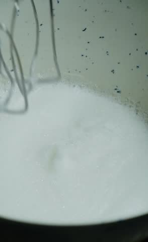 A hand mixer mixes the egg whites and sugar. Vertical video of mixer blending with white sugar and egg ingredients for meringue. Food, home cooking dessert, sweet, equipment, and housework concepts.