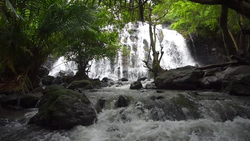 This waterfall is visible from a distance, partly covered by shady trees with lots of natural mossy rocks.