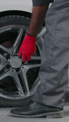 Medium vertical shot of legs and hands of anonymous dark-skinned male mechanic in gray overalls, red gloves tightening bolts on wheel of clients car with manual wrench after tire repair