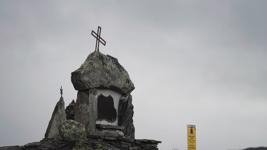 A monument with a metal crosse stands atop a rocky formation, set against a cloudy sky in the mountains. Parallax shot.