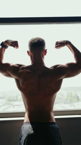 Fit man flexing his back muscles and posing in a gym. Strong physique and fitness training showcase strength and dedication. Large sunlit window in the background adds to the atmosphere. Vertical